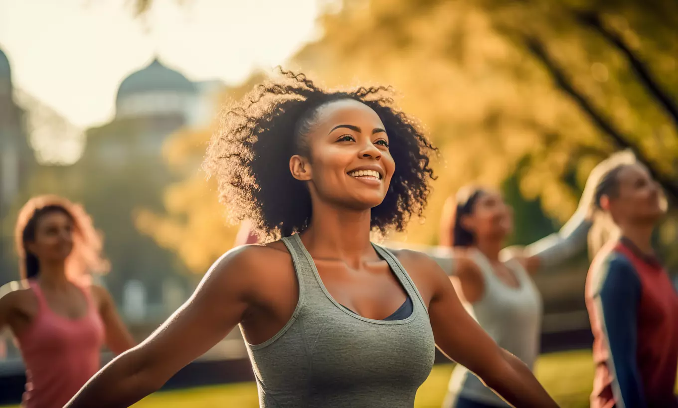 A beautiful black woman smiling and participating in yoga outside with others blurred in the background.