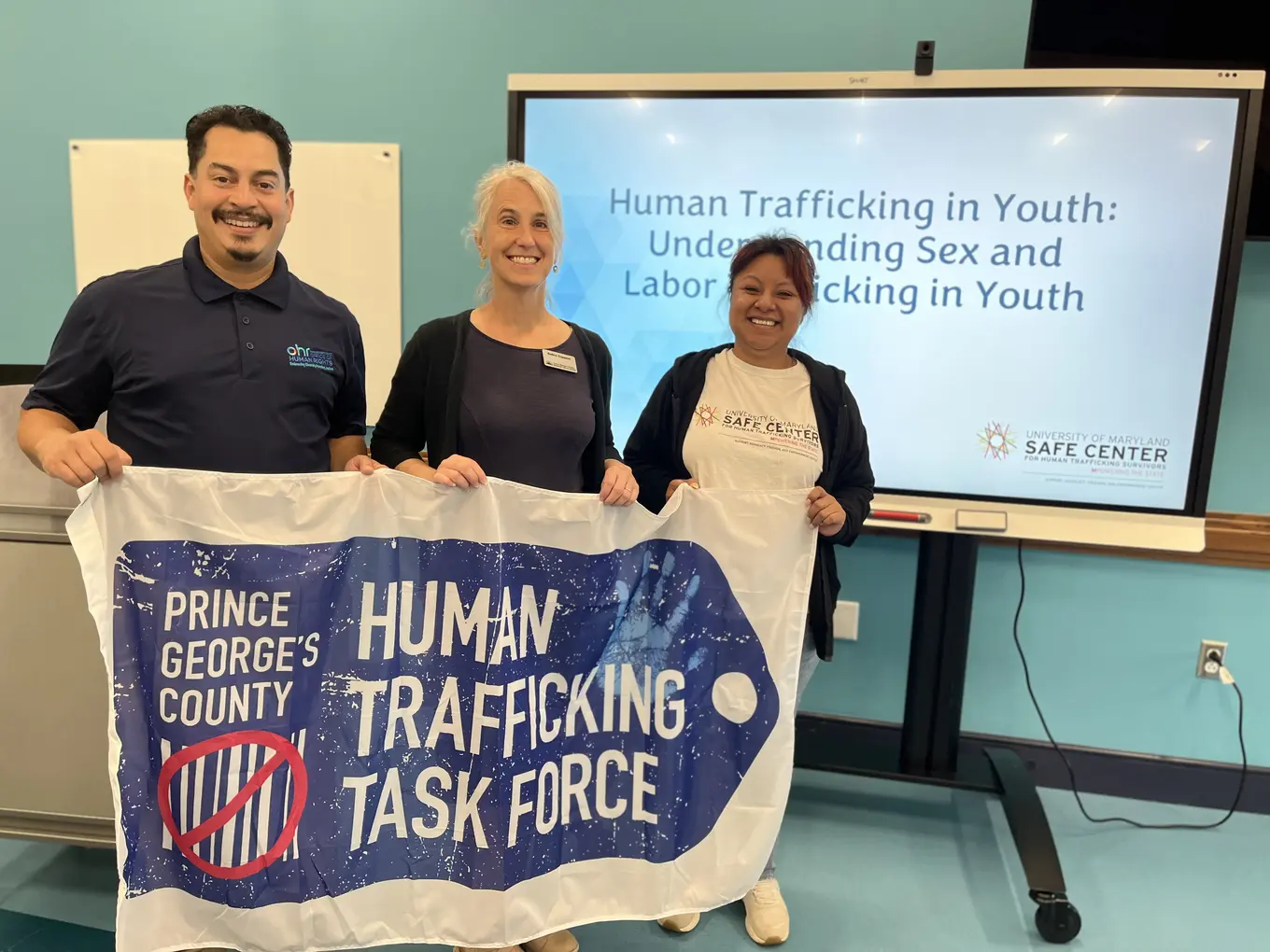 Three people holding a human trafficking task force banner standing in front of a screen
