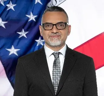 Image of Jose Villegas, a man with short hair wearing glasses, with an American flag in the background