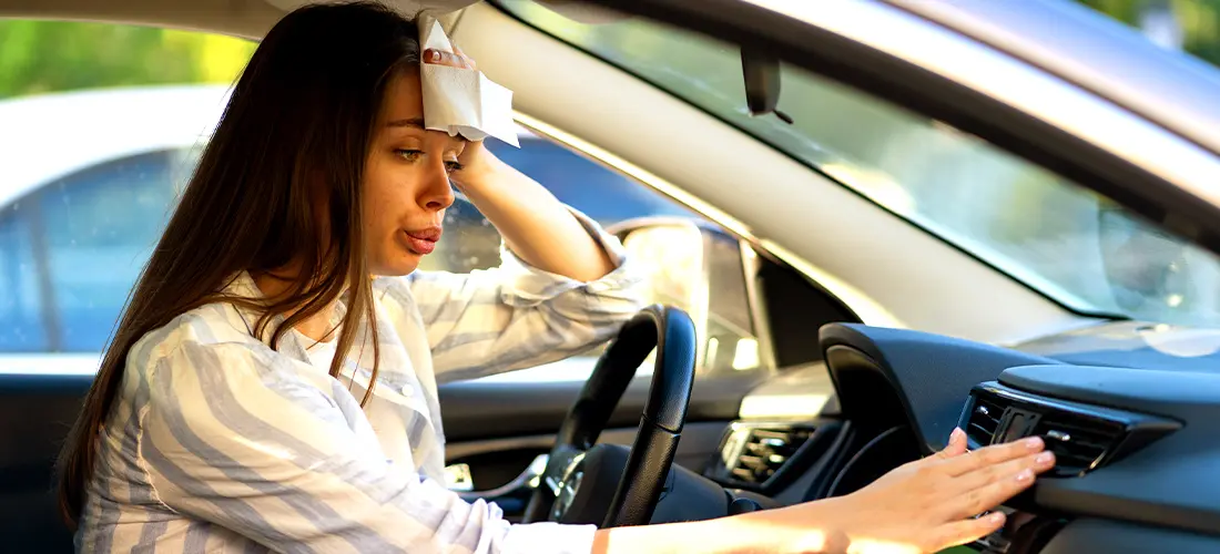 A woman frantically trying to cool down her car with the AC, wiping the sweat off her brow with a napkin in the extreme heat.